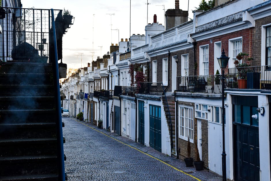 A narrow cobbled street lined with a row of terraced houses featuring white and brick facades, some with decorative white molding and window frames. The houses have small front gardens and balconies with black metal railings, some adorned with potted plants and flowers. To the left, a black staircase leads up to an unseen upper level. The street is illuminated by soft, ambient light, suggesting late afternoon or early evening. The scene showcases typical London-style architecture with chimneys visible on rooftops, and a few cars are parked along the street. The overall setting appears quiet and well-maintained, reflecting the charm of residential outdoor environments in the N1 area near Angel Station, with a focus on the exterior features of the buildings and the textured cobblestone street. This image is related to garden and outdoor space maintenance, as highlighted by Gardeners Islington, specialists in garden care and landscaping for terraces and properties in the area.
